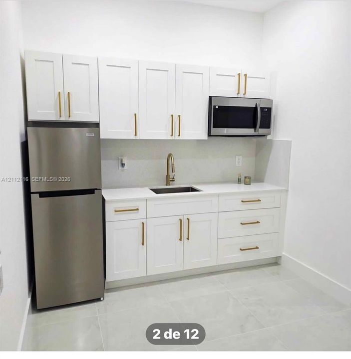 White shaker-style cabinetry with gold-toned hardware, quartz countertops, stainless steel refrigerator and built-in microwave, under-mount sink with gold-toned faucet, and light-toned tile flooring