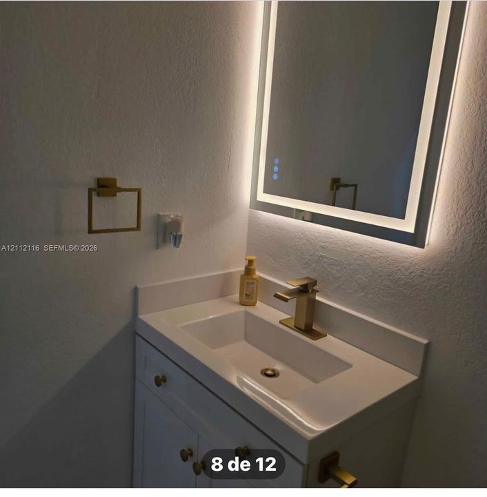 White vanity with an integrated basin, quartz-finish countertop, backlit LED mirror, and gold-toned hardware and fixtures
