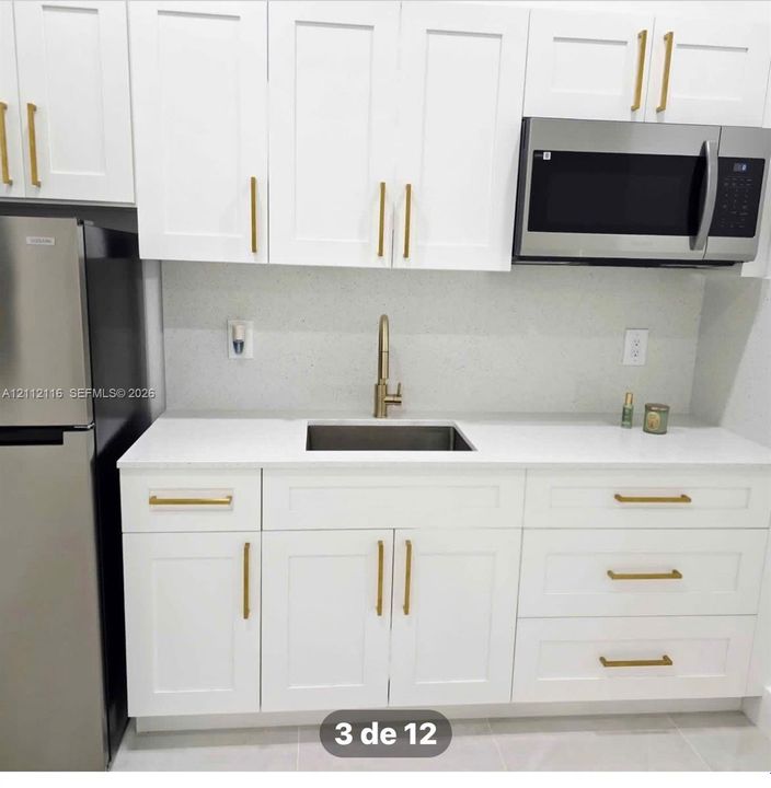 White shaker-style cabinetry with gold-tone hardware, solid-surface countertops, undermount stainless steel sink, gold-tone faucet, and built-in stainless steel microwave