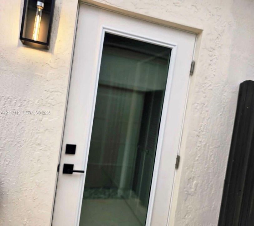 Textured stucco exterior featuring a full-light glass door, matte black hardware with electronic deadbolt, and a rectangular wall-mounted lantern with a filament bulb