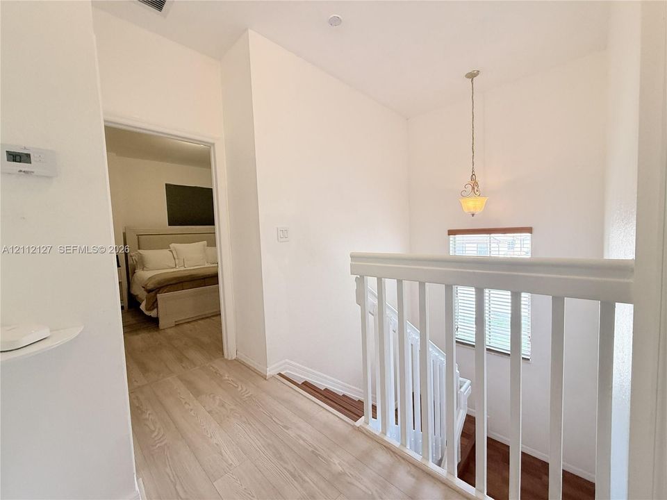 Hallway featuring wood-finish flooring, white spindle railing, a pendant light fixture, and horizontal window blinds
