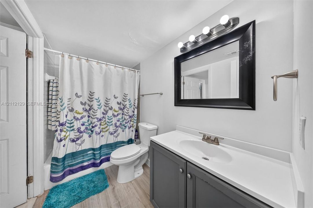 Bathroom featuring a gray shaker-style vanity with an integrated white sink, a black-framed wall mirror, multi-bulb vanity lighting, and wood-finish flooring