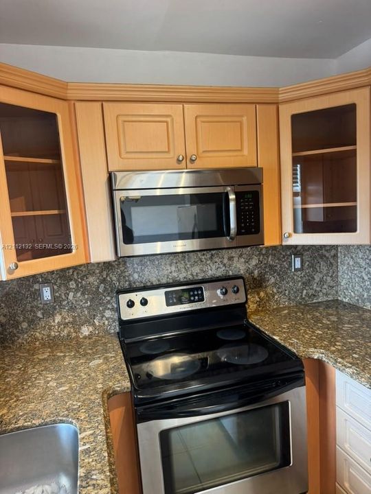 Kitchen featuring light-tone cabinetry with glass-front display panels, stone-surface countertops, stone backsplash, and stainless-steel appliances