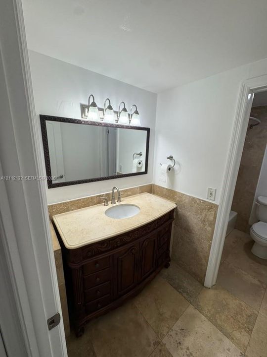 Dark wood vanity with an integrated stone countertop, undermount sink, framed rectangular wall mirror, four-light vanity fixture, and stone tile flooring
