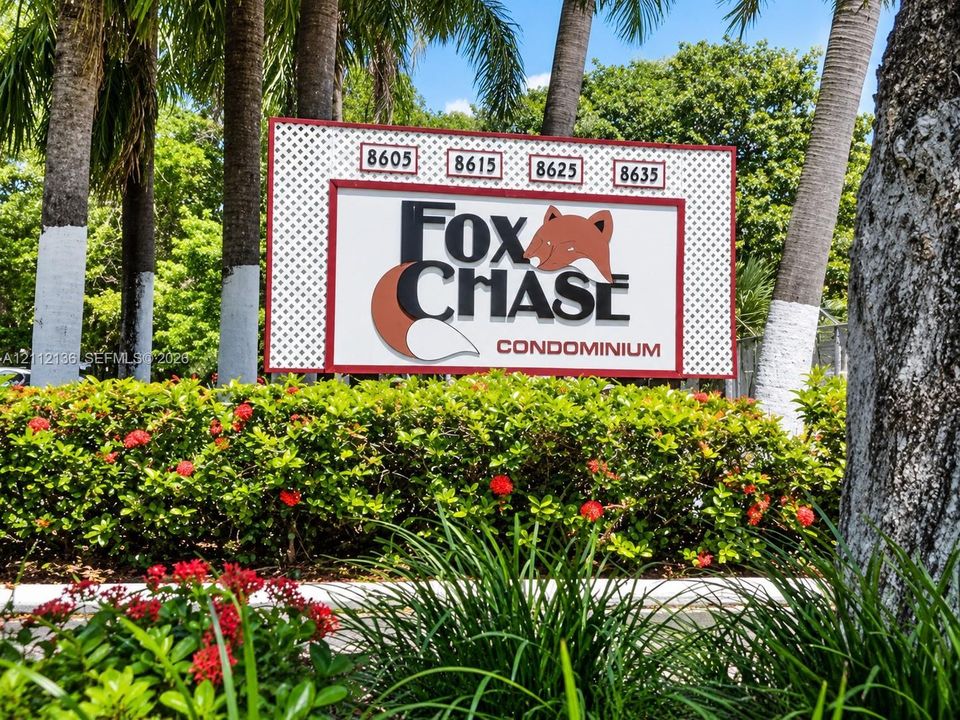 Community entrance signage for Fox Chase Condominium featuring mature palm trees and manicured shrubbery with flowering plants