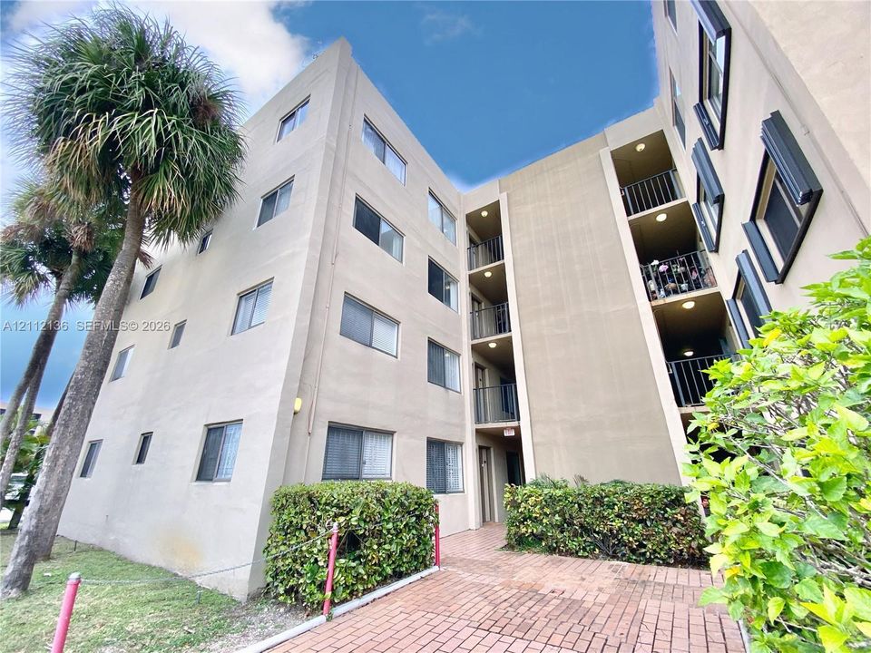 Multi-story exterior featuring a stucco facade, stacked balconies with metal railings, a brick-paved entrance path, and mature palm trees