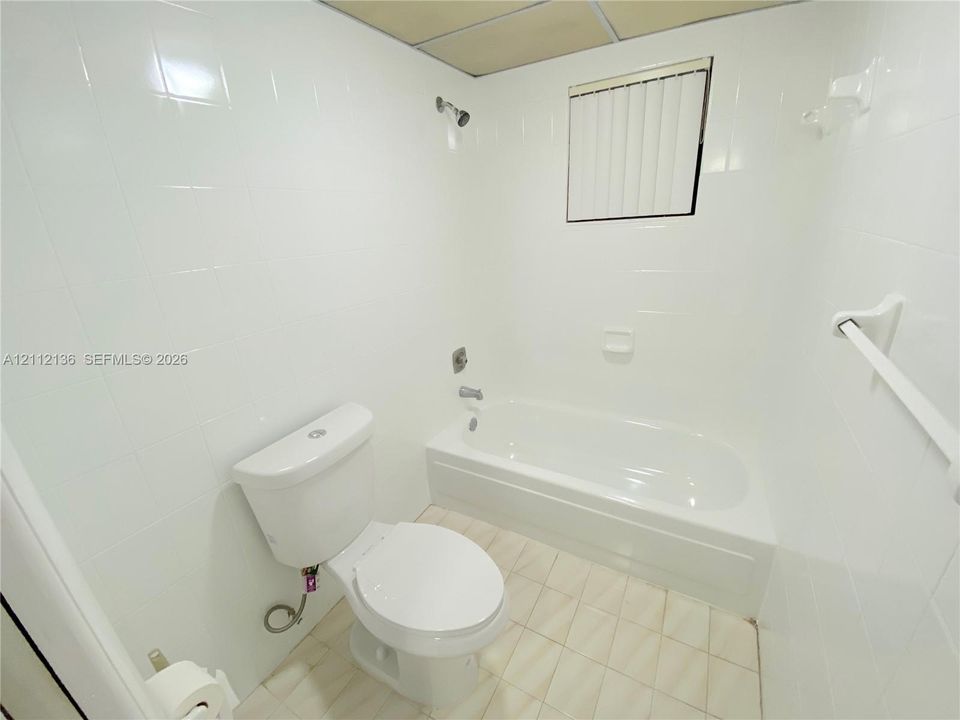 Full-height white wall tiling, porcelain bathtub with integrated shower head and diverter, porcelain toilet, and grid-pattern tile flooring