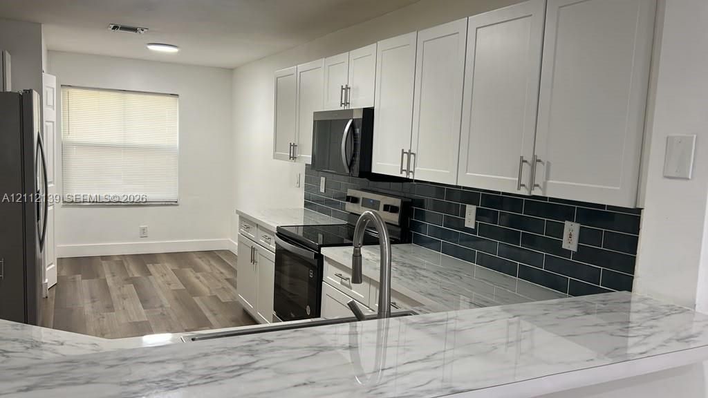 Kitchen featuring white shaker-style cabinetry, a dark tile backsplash, stone-finish countertops, stainless steel appliances, and wood-finish flooring