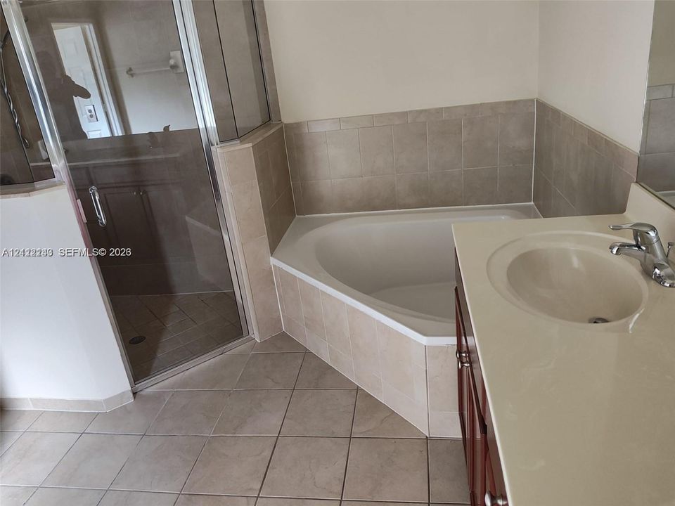Corner soaking tub with tile surround, glass-enclosed shower, wood-finish cabinetry, and solid-surface countertop with integrated sink