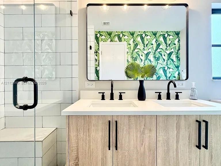 Dual-sink vanity with light-tone wood-finish cabinetry, white countertop, rectangular undermount sinks, and matte black fixtures, paired with a walk-in shower featuring subway tile surrounds and a glass enclosure
