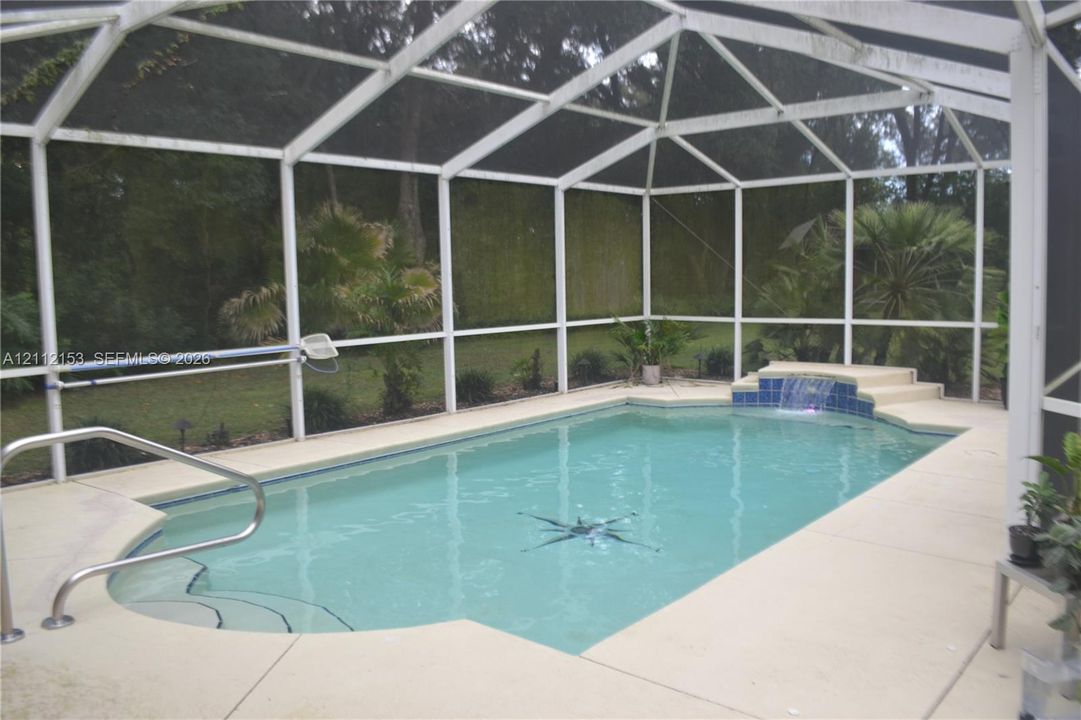 Screened-in pool area featuring a built-in water feature, concrete decking, metal pool handrails, and an inlaid pool floor medallion