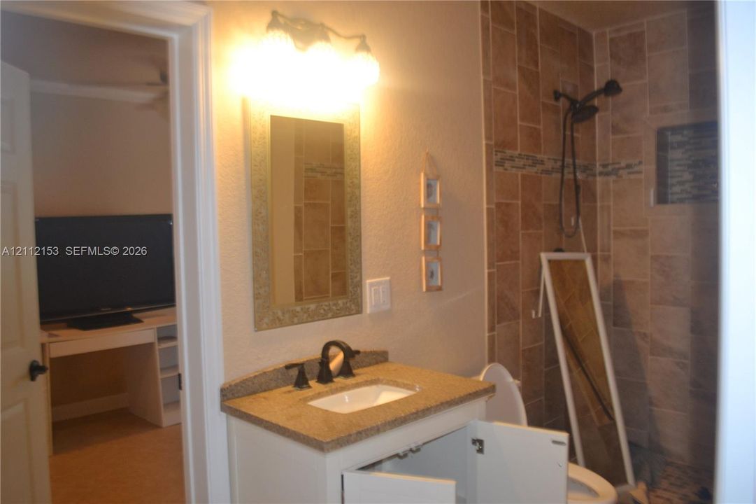 Bathroom vanity with stone-finish countertop, undermount sink, and oil-rubbed bronze faucet, featuring a wall-mounted light fixture, framed mirror, and walk-in shower with tile surround