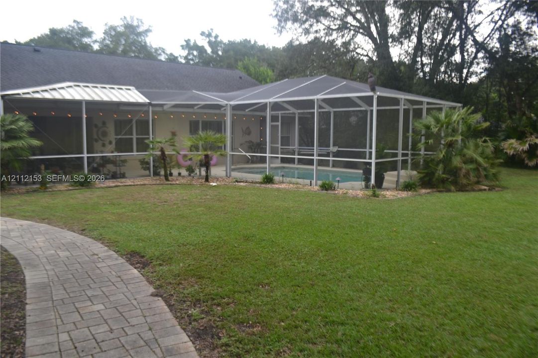 Screen-enclosed swimming pool featuring a paver stone walkway, mature landscaping, and an expansive lawn