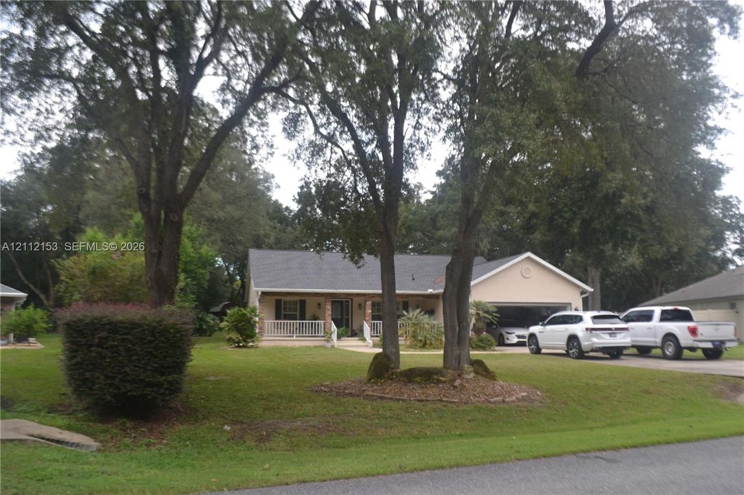 Single-story exterior featuring a covered front porch, light-toned stucco facade, dual-car garage, and mature shade trees