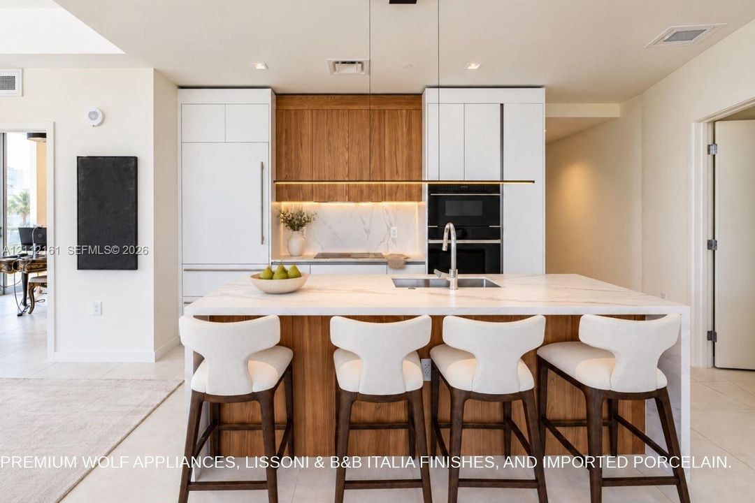 Kitchen featuring an expansive center island with a stone-finish countertop, integrated built-in appliances, flat-panel cabinetry in white and natural wood finishes, and under-cabinet LED task lighting