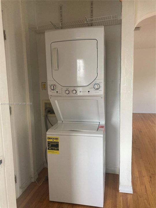 Stacked white laundry center with integrated dryer and top-loading washer, housed in a dedicated alcove featuring white wire shelving and wood-finish flooring