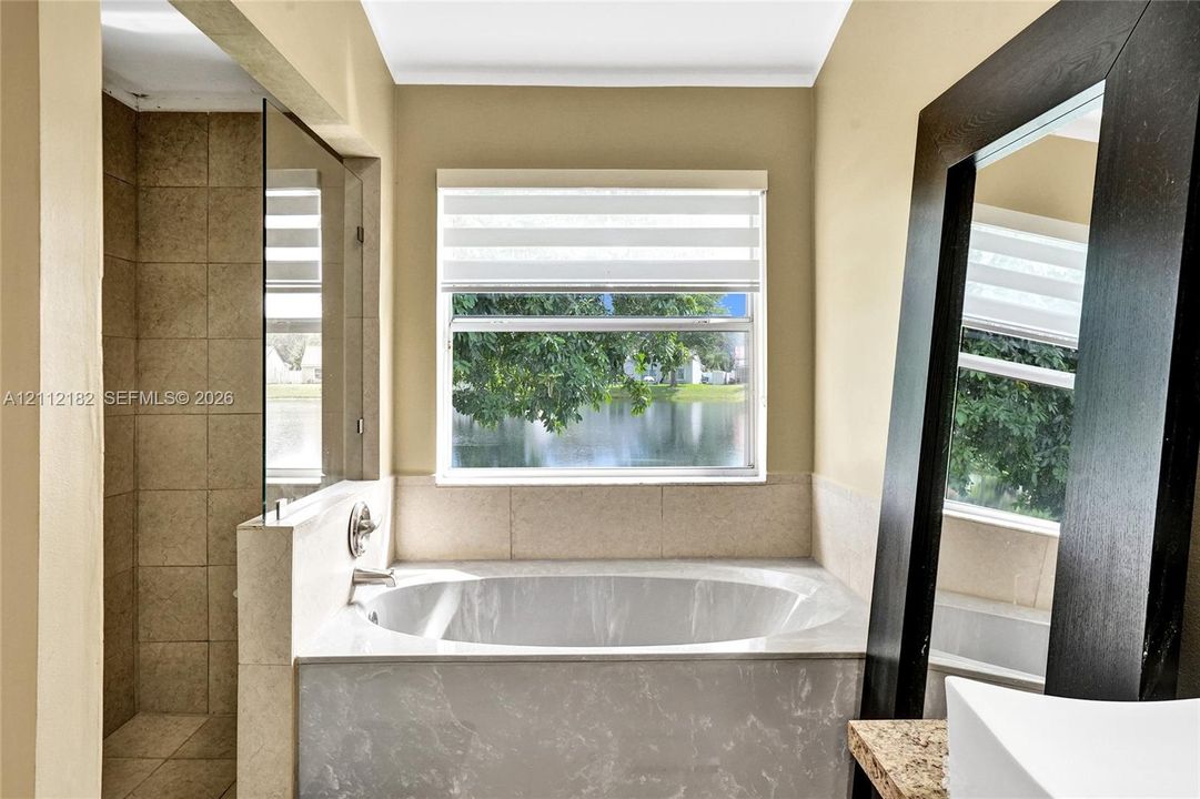 Soaking tub with a stone-finish surround, walk-in shower with floor-to-ceiling tile, and large window with exterior water backdrop