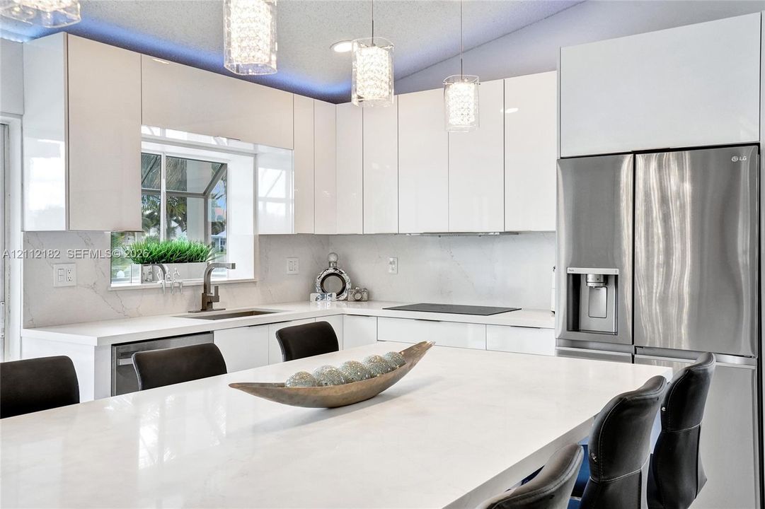 Kitchen featuring white high-gloss cabinetry, stone countertops with matching backsplash, central island, stainless steel refrigerator, and pendant lighting
