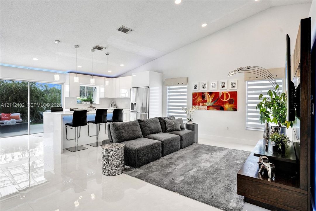 Open-concept interior featuring vaulted ceilings, recessed lighting, white cabinetry with integrated appliances, a kitchen island with stone-finish countertops, and polished tile flooring