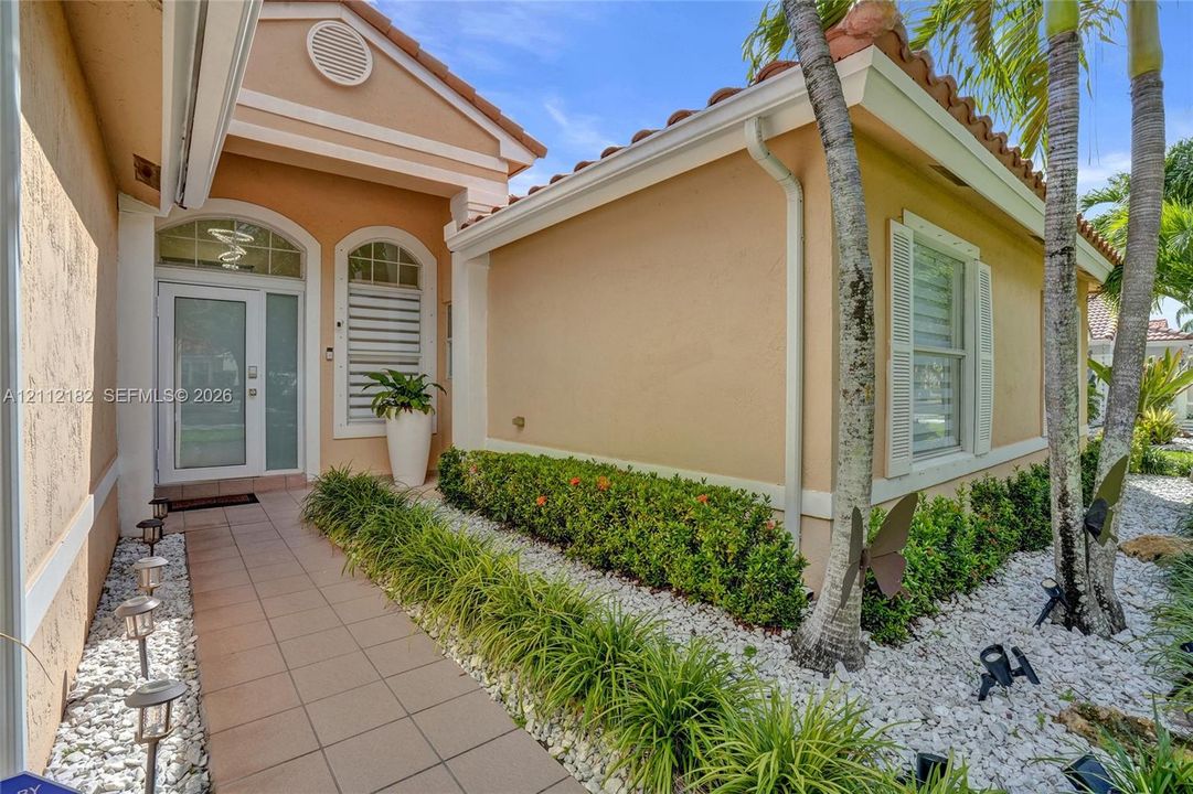 Stucco exterior with tile roof, tiled entryway, glass-paneled front door with arched transom window, and landscaped stone garden beds