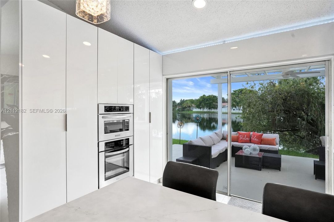 Floor-to-ceiling high-gloss cabinetry featuring an integrated stainless steel double oven and microwave, stone-finish island, and large glass sliding doors opening to a covered patio with water frontage