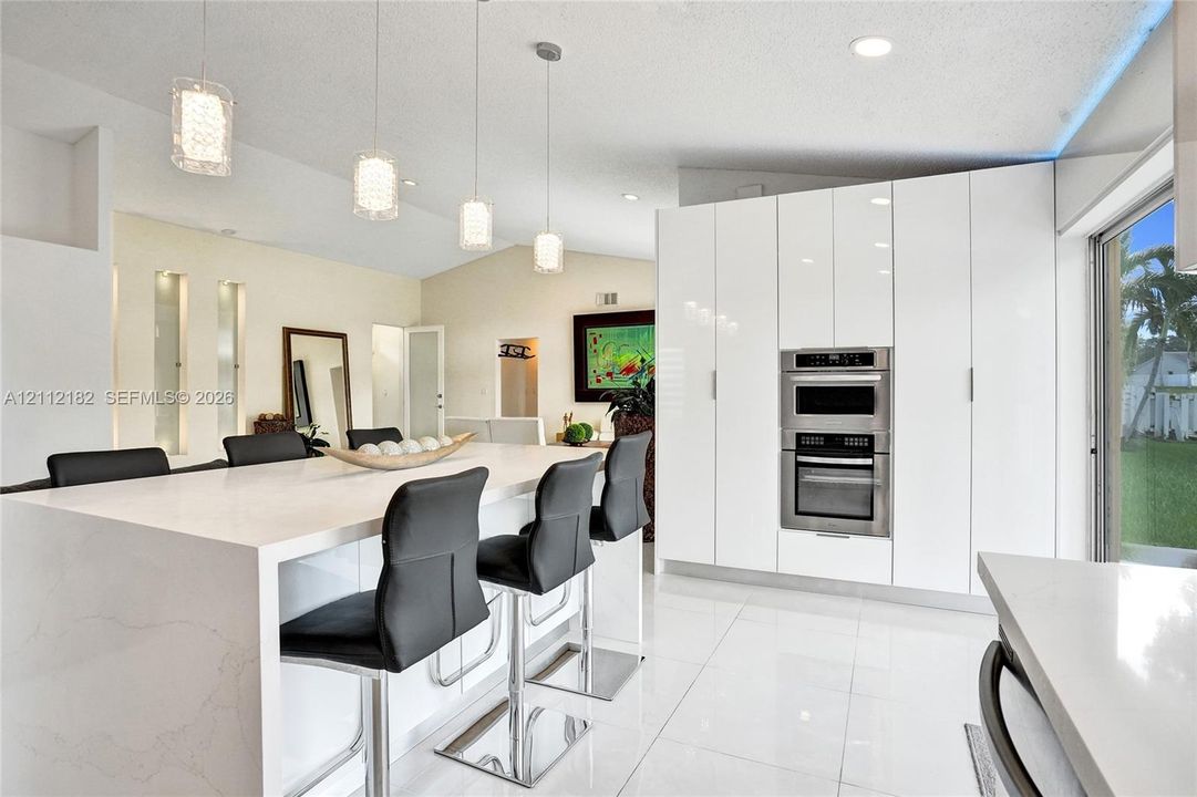 Modern kitchen featuring white flat-panel cabinetry, a central island with a solid surface countertop, built-in stainless steel double ovens, tile flooring, and multi-light pendant fixtures