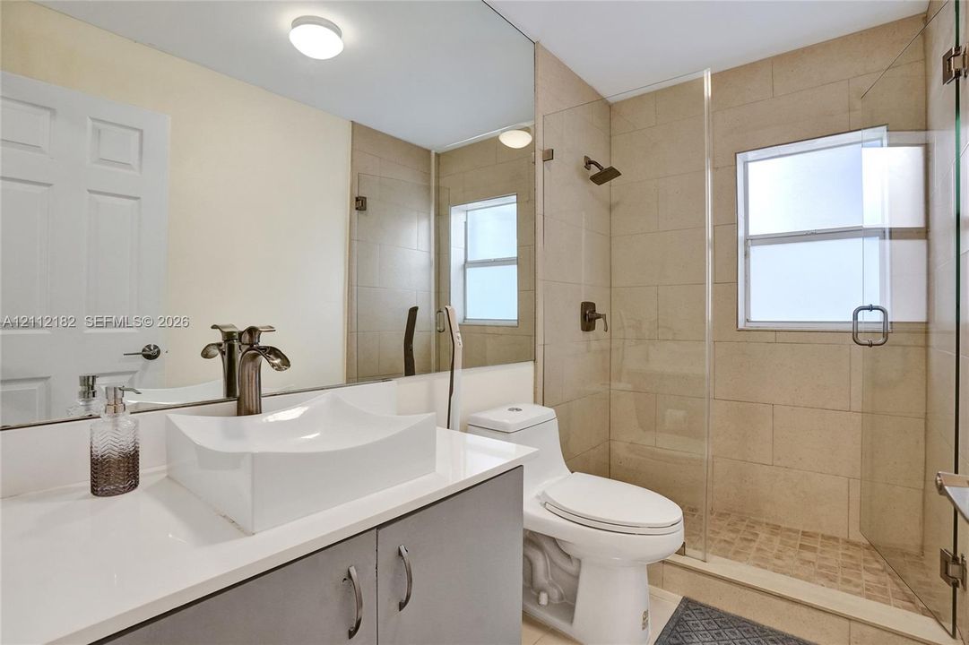 Bathroom featuring a vessel sink with a dark metal faucet, expansive vanity mirror, walk-in shower with floor-to-ceiling neutral tiling, and glass enclosure