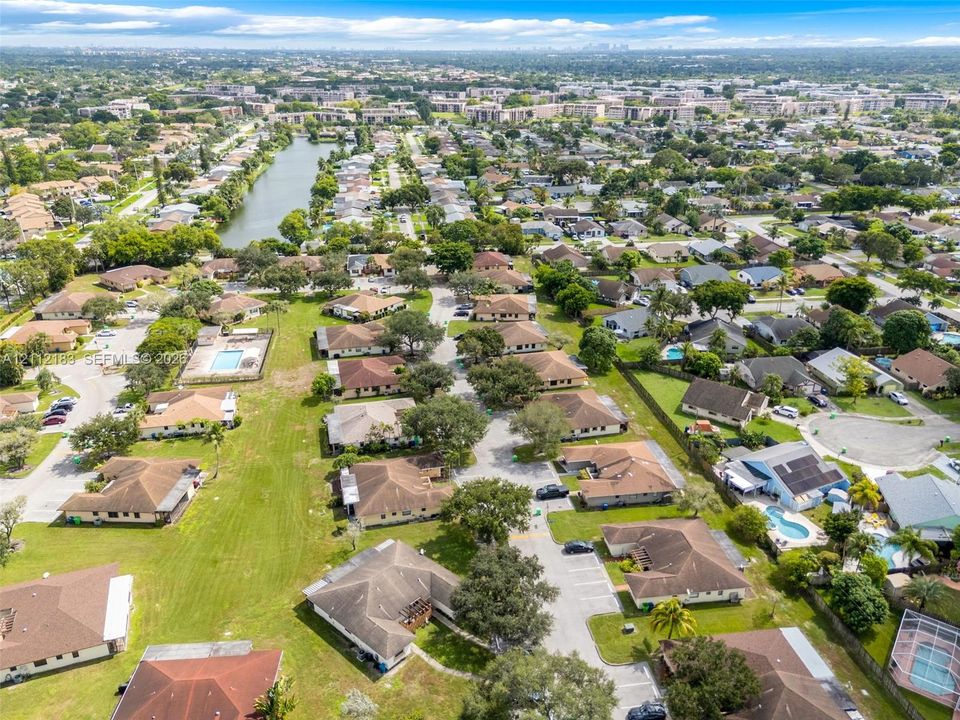 Aerial perspective of a suburban community featuring a centralized lake, mature landscaping, paved roadways, and residential properties with tiled roofs