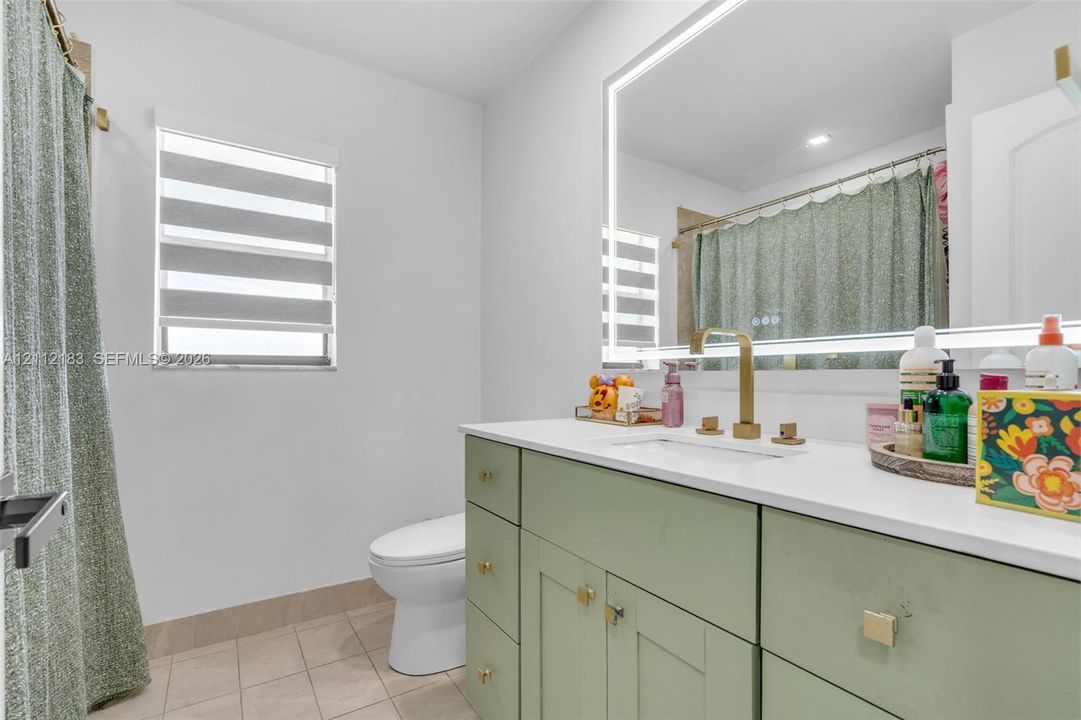 Bathroom featuring a green-toned shaker-style vanity, white surface countertop, integrated sink, brass-finish widespread faucet, and a large backlit rectangular mirror