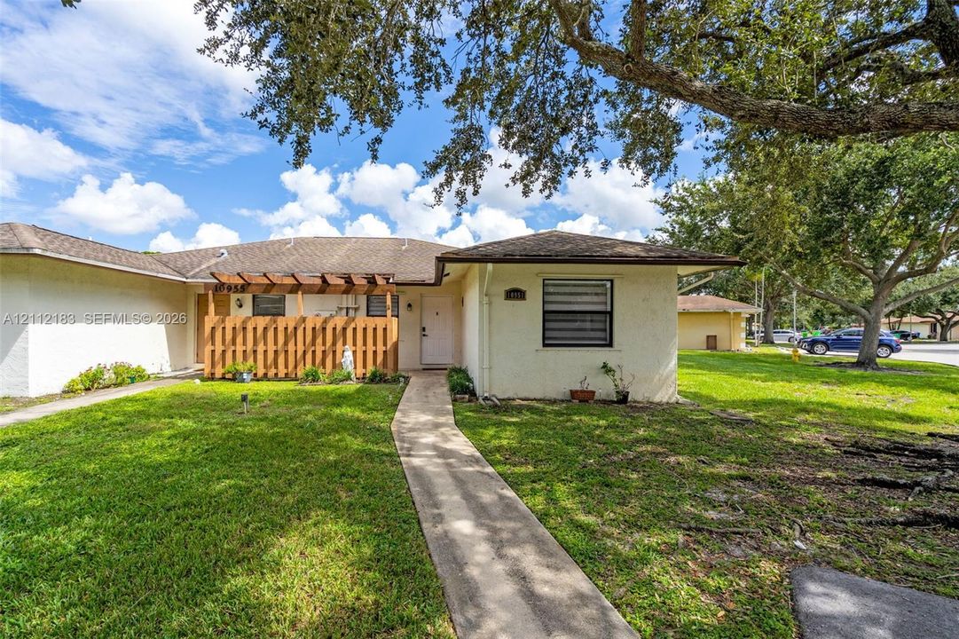 Stucco exterior with a hip roof, featuring an attached wood pergola, concrete entryway walkway, and mature trees