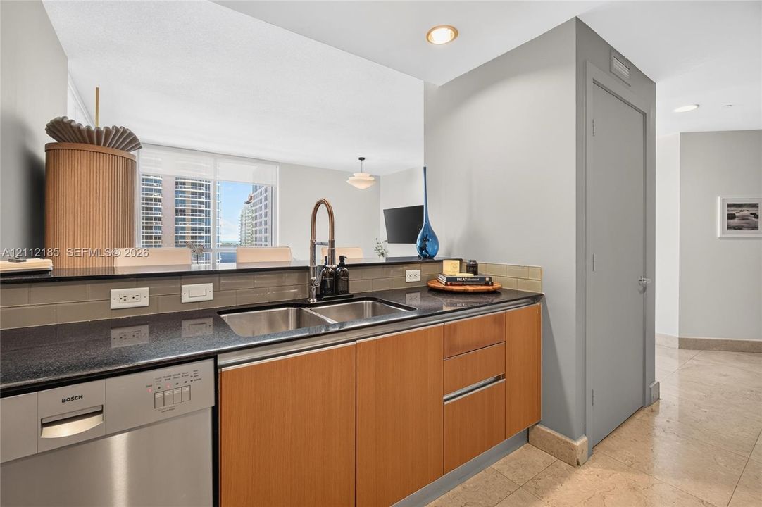 Kitchen area featuring stone countertops, natural wood-finish cabinetry, stainless steel dishwasher, undermount dual-basin sink with pull-down faucet, and neutral subway tile backsplash