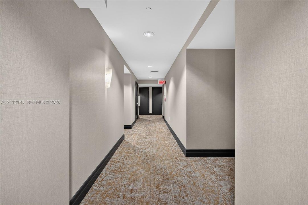 Hallway featuring textured neutral wall covering, patterned carpeted flooring, contrasting dark trim, and wall-mounted sconce lighting