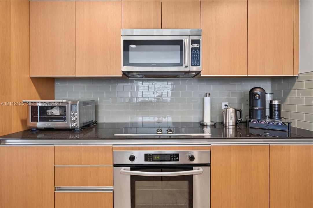 Kitchen featuring light wood-finish flat-panel cabinetry, dark stone countertops, stainless steel built-in oven, integrated microwave, and a glass subway tile backsplash