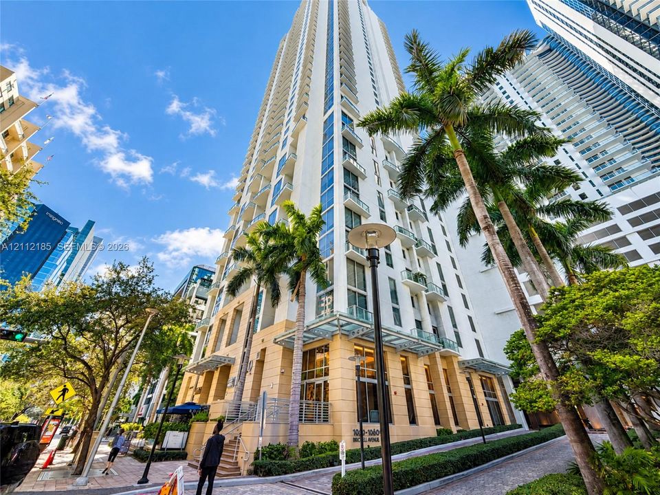 High-rise building exterior featuring a light-toned masonry facade, floor-to-ceiling glass paneling, stacked private balconies, and glass-canopy entryway detailing
