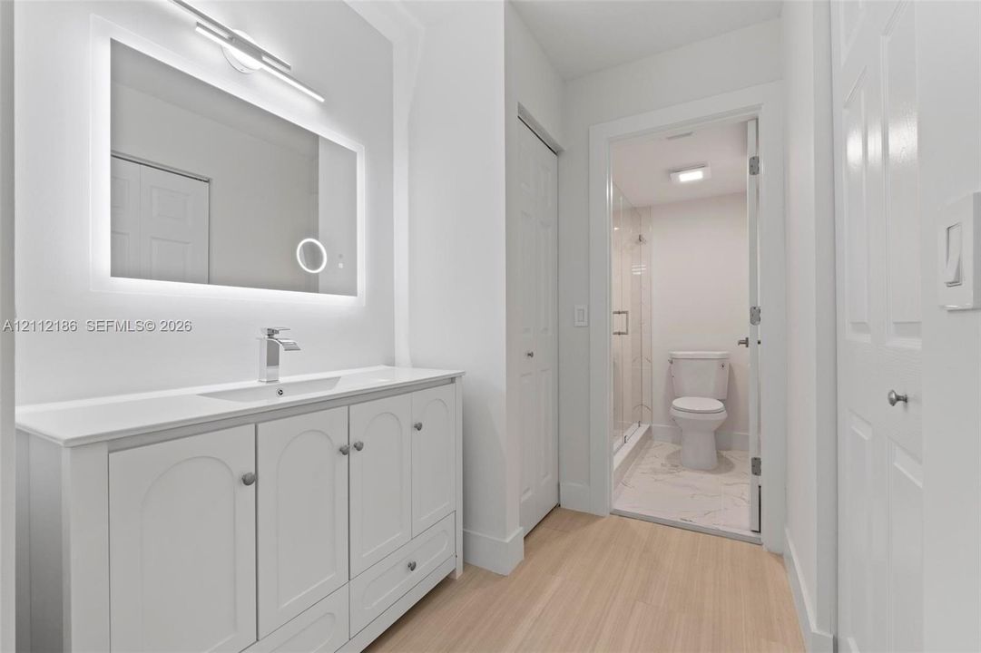 White vanity with integrated sink, backlit LED mirror, wood-finish flooring, and walk-in shower with glass enclosure