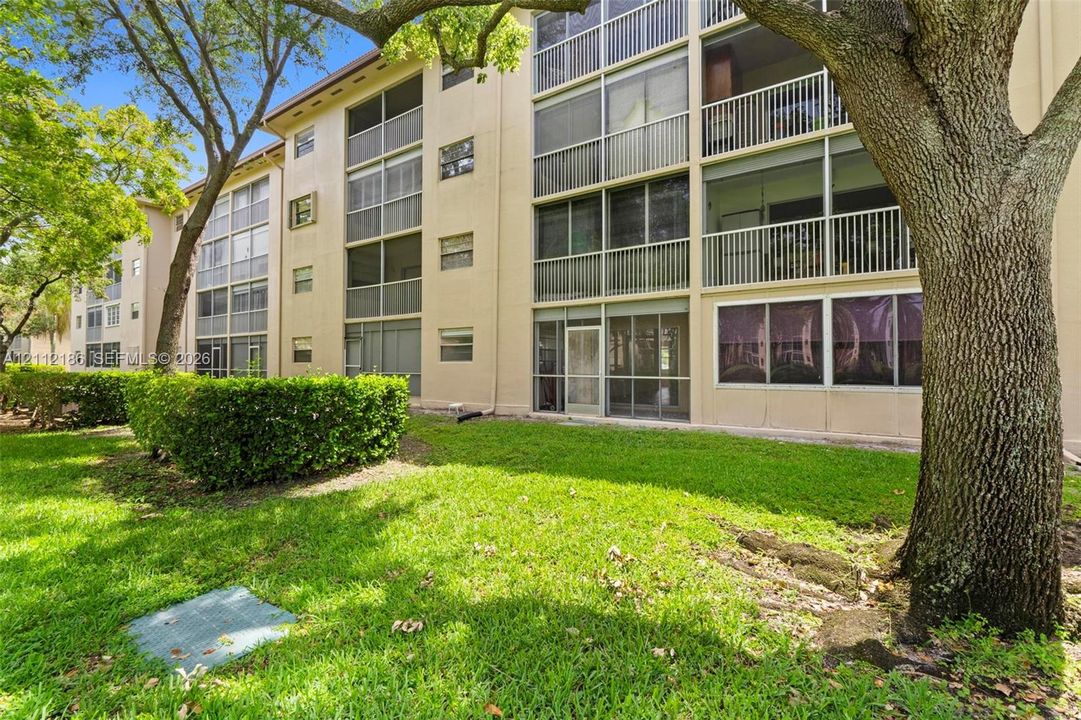Multi-story building exterior featuring screened-in lanais, vertical sliding windows, light-toned stucco facade, and manicured landscaping with mature trees