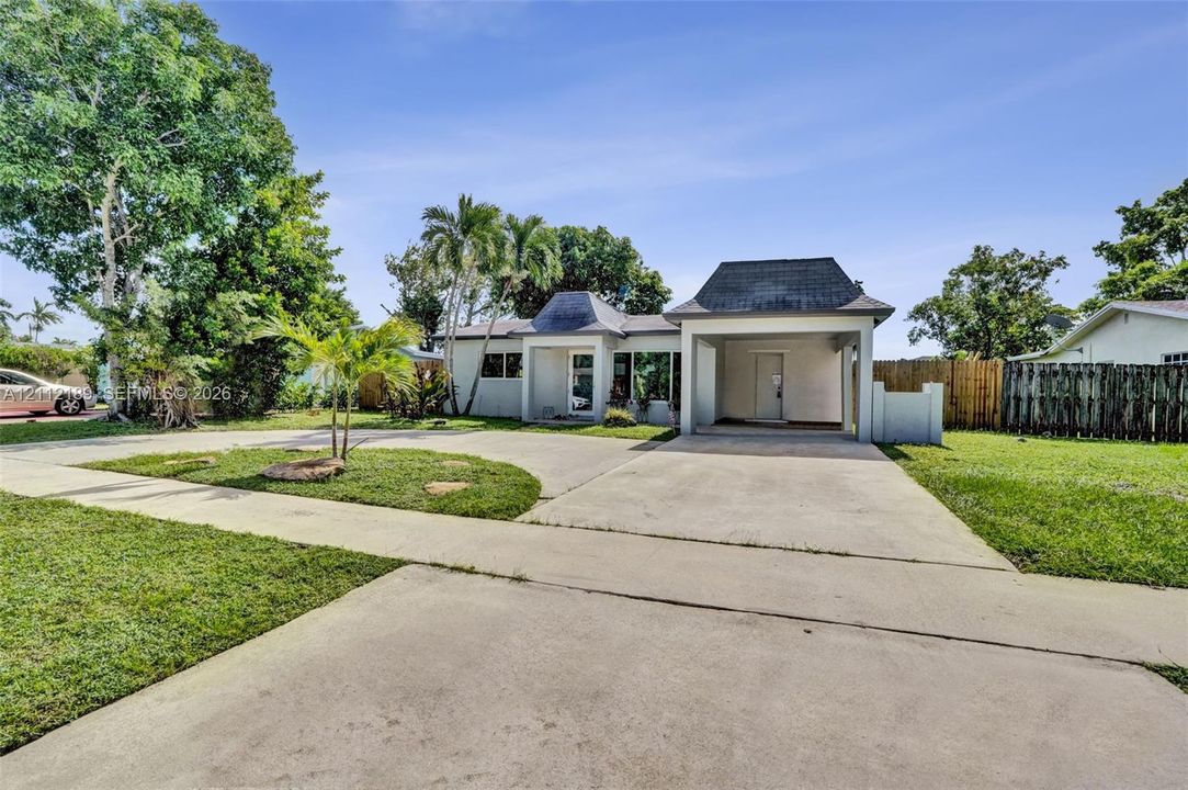 Single-story residence featuring a covered carport, pitched shingle roofline, concrete driveway, and expansive lawn with mature palm trees
