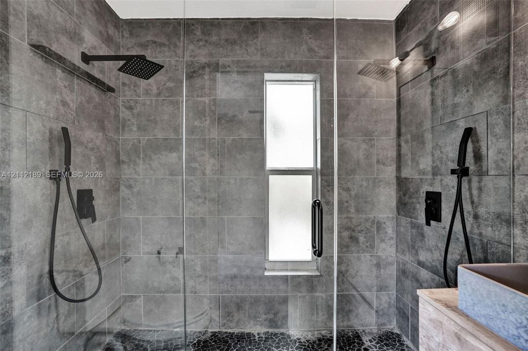 Walk-in shower featuring large-format grey stone wall tile, black matte rainfall showerheads, handheld shower fixtures, a glass door enclosure, and a stone-tiled floor