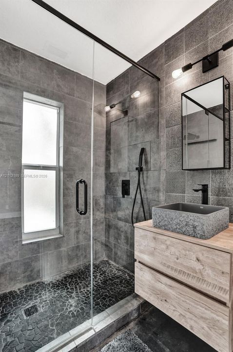Walk-in shower featuring glass paneling, floor-to-ceiling gray tile, and a pebble-stone basin