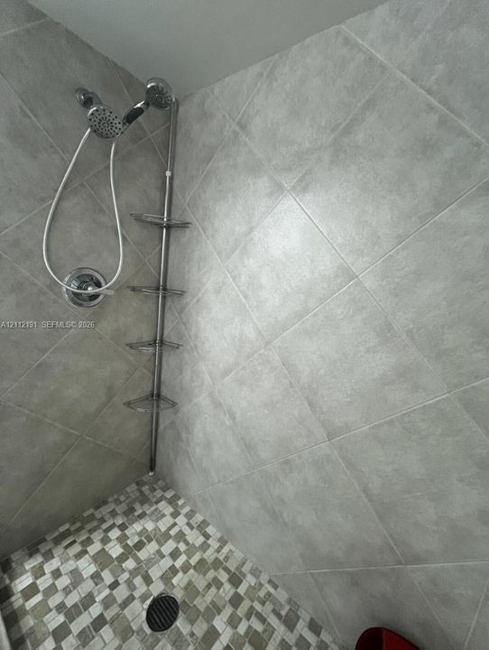 Shower enclosure featuring large-format diagonal wall tiling, mosaic floor tile, multi-setting handheld shower head, and a vertical stainless steel corner shelving unit
