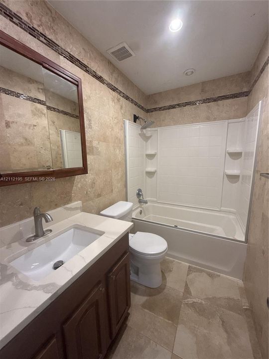 Bathroom featuring stone-finish wall and floor tiling, a vanity with a white countertop and rectangular undermount sink, a framed medicine cabinet, and a bathtub with a white surround