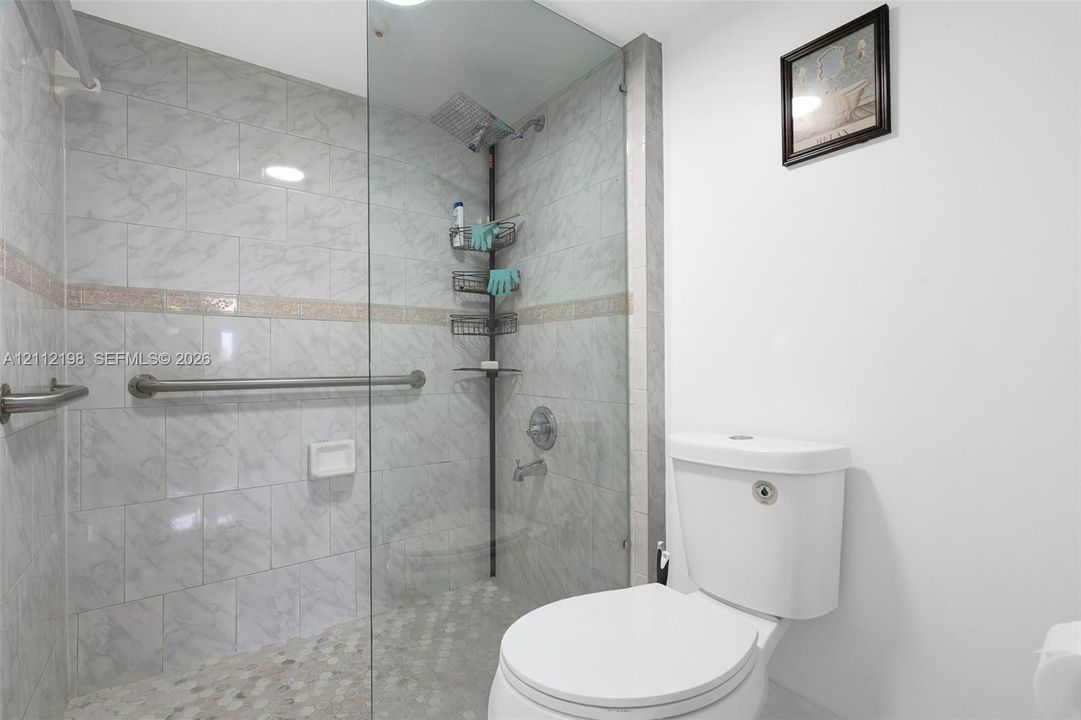 Walk-in shower with floor-to-ceiling stone-patterned tile, glass partition, wall-mounted metal grab bar, and rain-style showerhead, complete with a dual-flush toilet