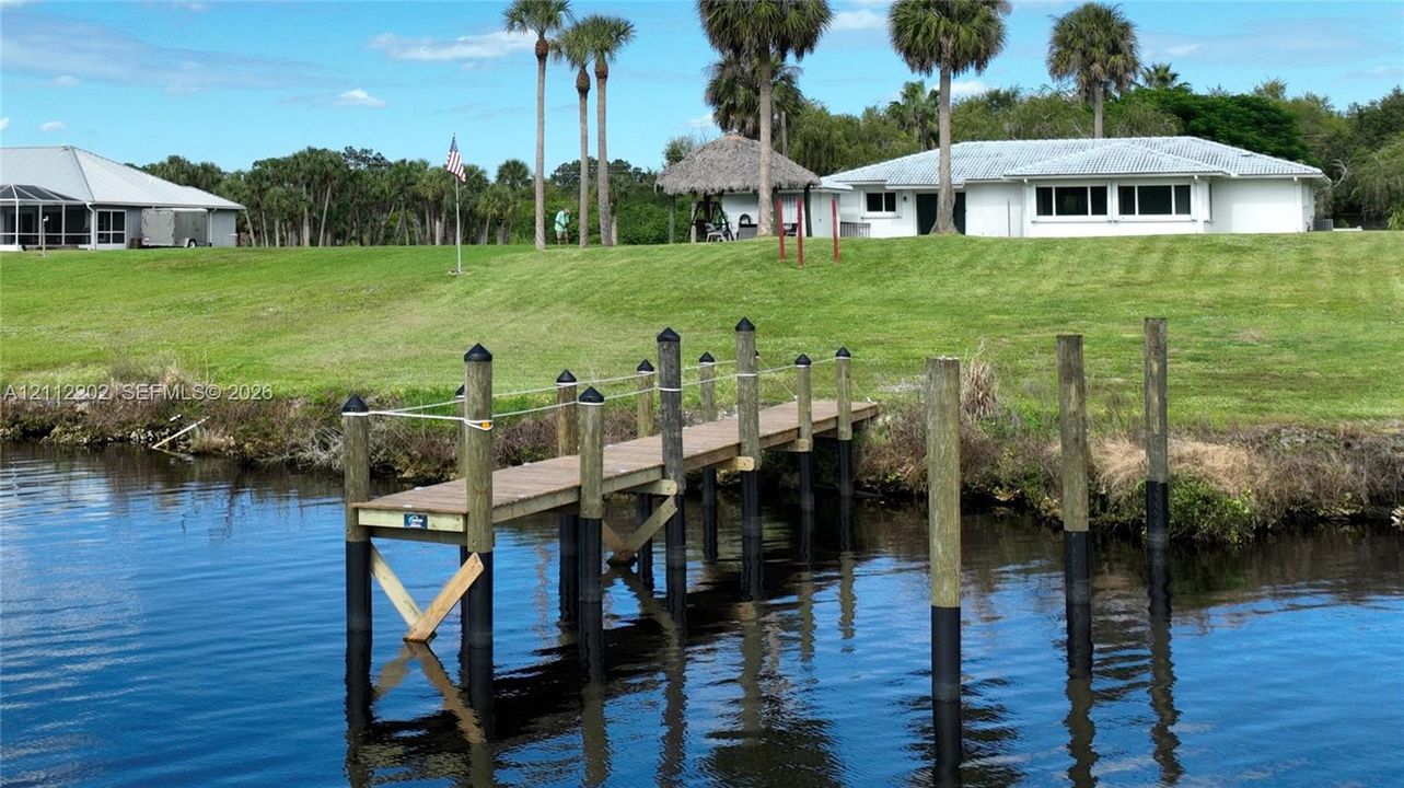 Waterfront property featuring a private wooden dock, sprawling grass grounds, mature palm trees, and a detached thatched-roof structure