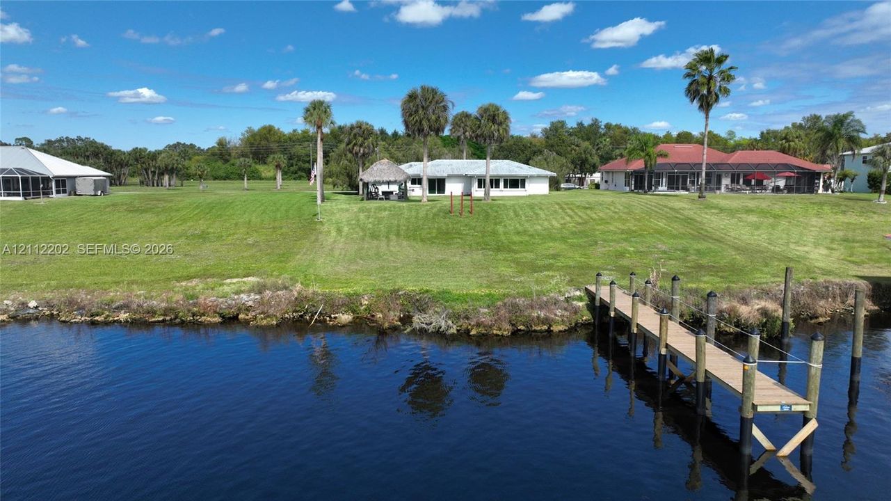 Waterfront property featuring a private wooden dock, expansive lawn, detached thatched-roof structure, and mature palm trees