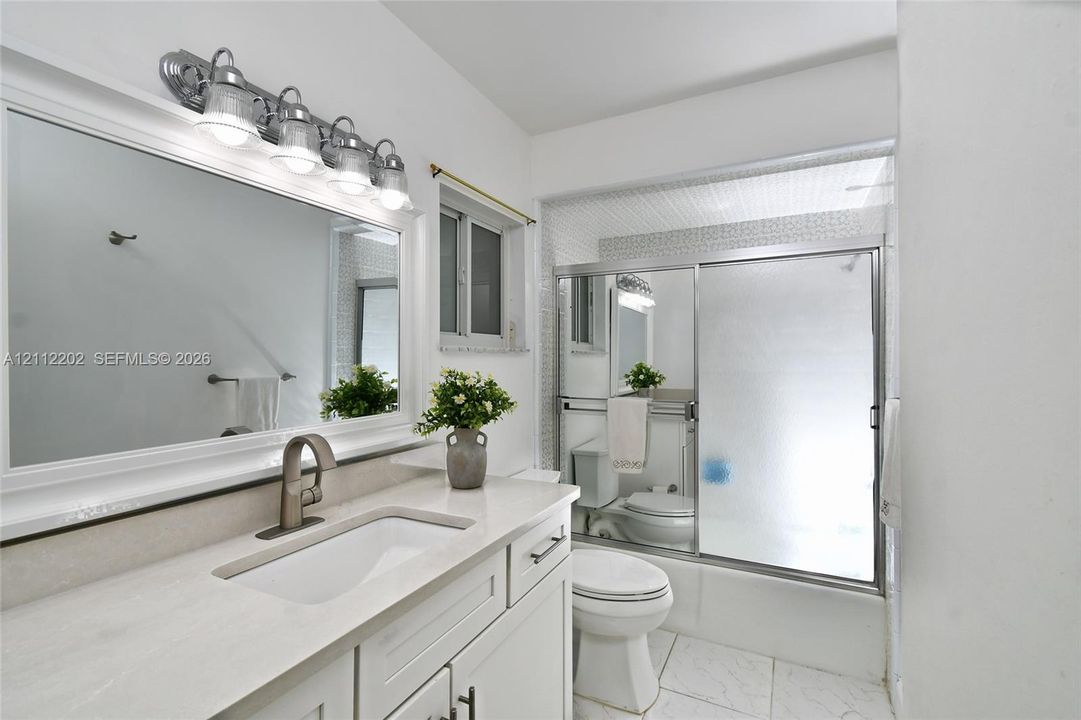 Bathroom featuring a white vanity with a composite countertop, undermount sink, wide-frame mirror, four-light vanity fixture, and a shower-tub combination with sliding glass doors