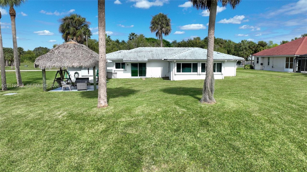 Rear exterior featuring a sprawling lawn, a detached thatched-roof palapa, and mature palm trees