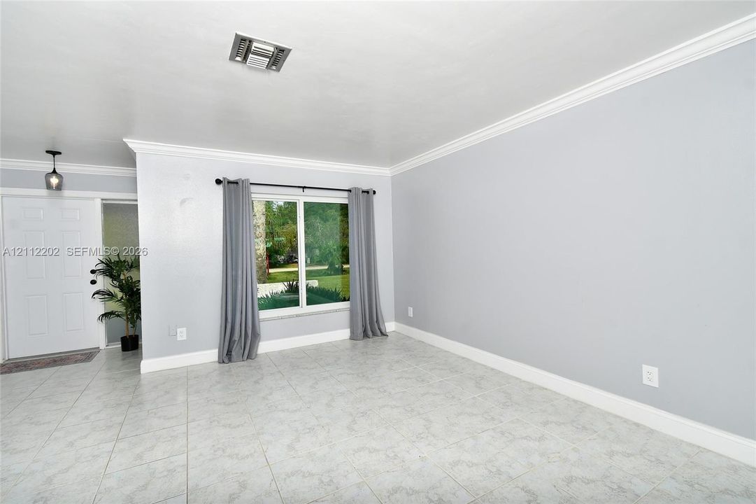 Living area featuring light-toned tile flooring, neutral wall paint, multi-pane window, crown molding, and a paneled entry door