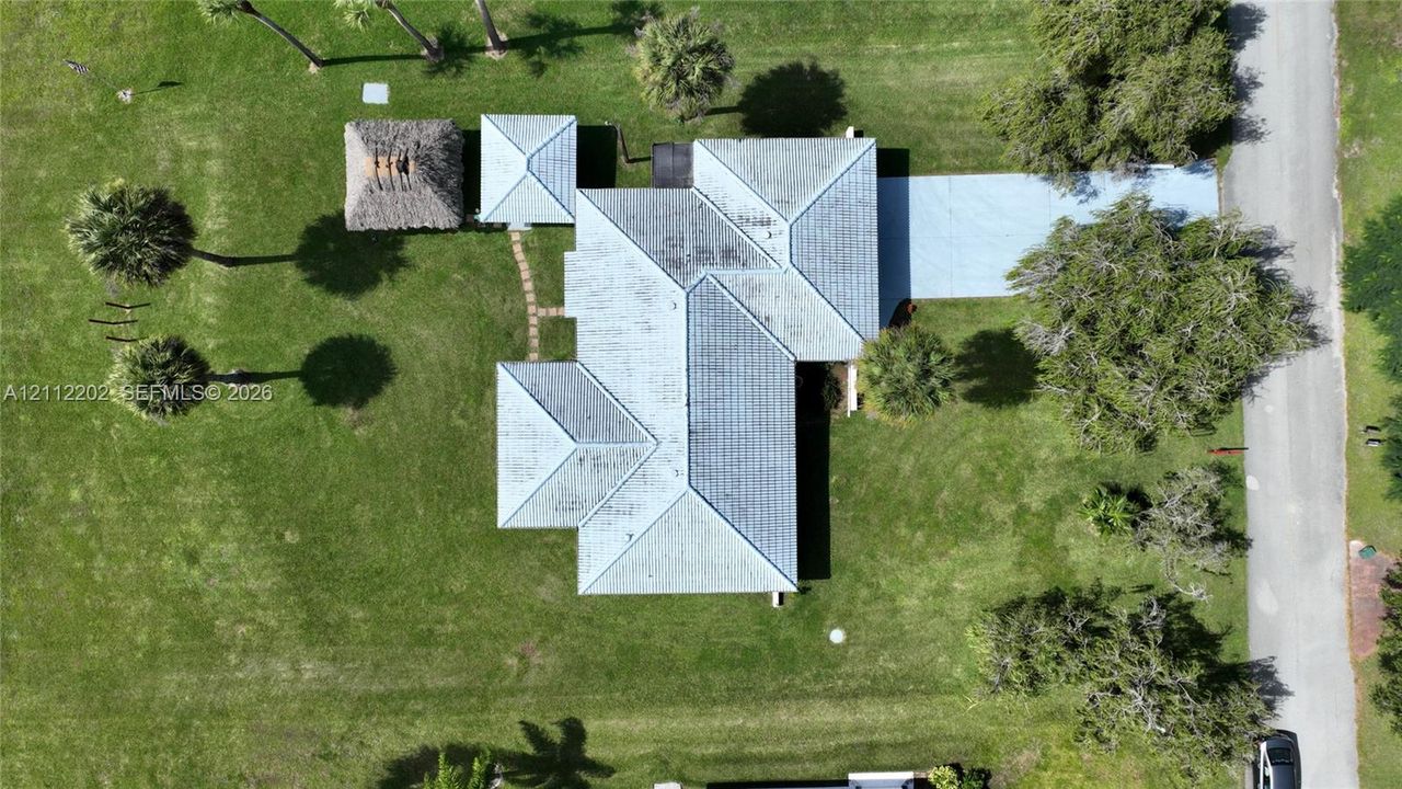 Aerial perspective of property featuring a multi-pitched metal roof, paved driveway, detached thatched-roof structure, and expansive grass lawn with mature palm trees