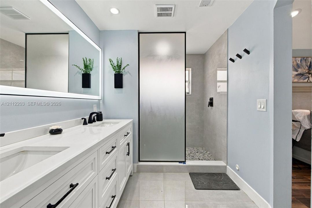 Double vanity with integrated sinks, quartz countertop, backlit mirror, matte black fixtures, and walk-in shower with frosted glass enclosure