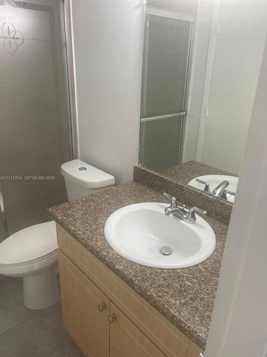 Bathroom vanity featuring a light wood-finish cabinet, stone-composite countertop, integrated porcelain sink, and a walk-in shower with a frosted glass door